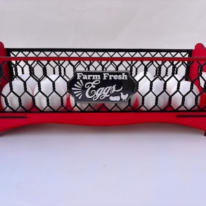 May include: A red and black wooden egg basket filled with white eggs. The basket has a decorative design and a sign that reads "Farm Fresh Eggs". The basket is designed to hold eggs.