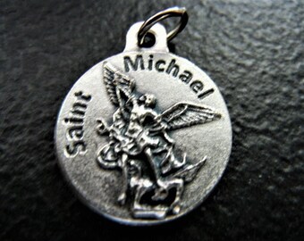 May include: Silver pendant with a depiction of Saint Michael the Archangel slaying a dragon. The pendant is inscribed with "Saint Michael" on the left side and "Michael" on the right side.
