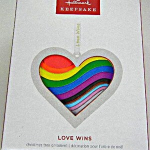 May include: A Hallmark Keepsake Christmas tree ornament box featuring a heart-shaped cutout revealing a rainbow-colored layered design. The box displays the text Hallmark KEEPSAKE, LOVE WINS, and christmas tree ornament in white and red.