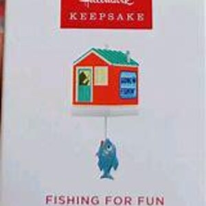 May include: A red and green Christmas ornament shaped like a small house with a blue fish hanging from it. The house has a sign that says "Gone Fishin'" and the fish has a white eye. The ornament is part of the Hallmark Keepsake collection and is titled "Fishing for Fun."