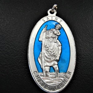 May include: Silver oval pendant with a blue enamel background. The pendant features a depiction of Saint Christopher carrying a child on his shoulders. The words "SAINT CHRISTOPHER" are inscribed around the edge of the pendant.