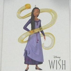 May include: A Hallmark Keepsake Christmas tree ornament box featuring Asha from Disney Wish. The box displays a graphic of Asha in a purple dress with a translucent yellow star trail swirling around her. Text includes Hallmark Keepsake, Disney WISH, and ASHA AND STAR.