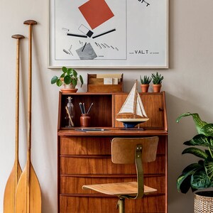 May include: A mid-century modern desk with a framed abstract art print, a small sailboat model, and potted plants. Two wooden oars lean against the wall. The desk is made of wood and has a matching chair.