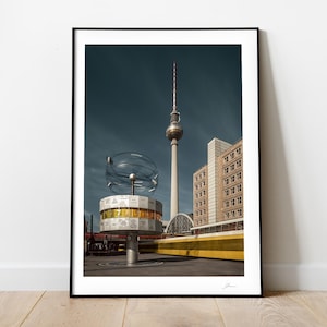 May include: A black and white photograph of the Berlin TV Tower, a tall, slender tower with a spherical observation deck, against a blue sky. In the foreground, a large, circular, metal sculpture with a clock face and a globe-like structure is visible.