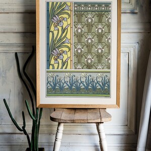 May include: A framed print of a vintage botanical illustration featuring a repeating pattern of stylized iris flowers in shades of green, yellow, blue, and white. The print is in a wooden frame and is sitting on a wooden stool.
