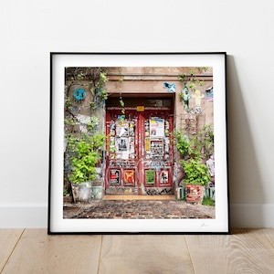 Pode incluir: Uma porta dupla vermelha com uma variedade de cartazes e adesivos cobrindo a superf&iacute;cie. A porta &eacute; cercada por plantas verdes e plantas em vasos. A porta est&aacute; localizada em uma parede de tijolo com uma pintura desbotada.