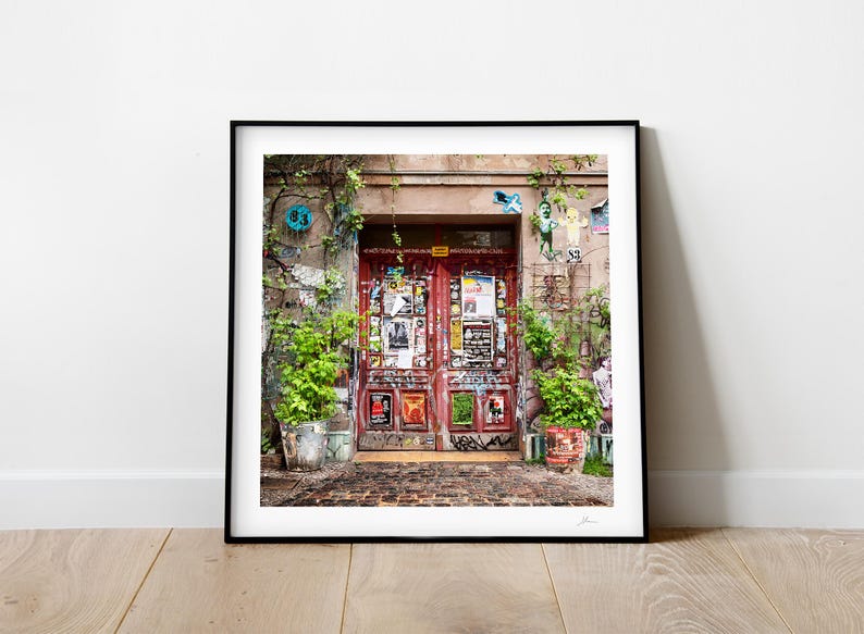 Pode incluir: Uma porta dupla vermelha com uma variedade de cartazes e adesivos cobrindo a superf&iacute;cie. A porta &eacute; cercada por plantas verdes e plantas em vasos. A porta est&aacute; localizada em uma parede de tijolo com uma pintura desbotada.