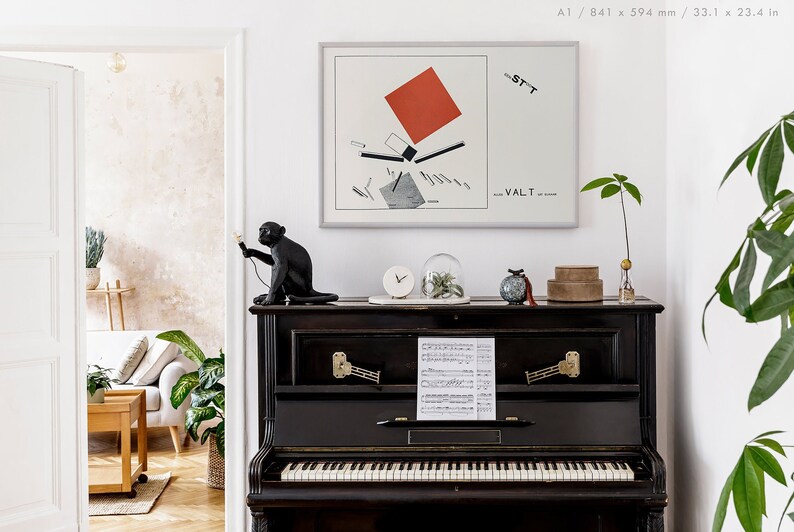 May include: A black upright piano with a framed abstract art print and decorative items. A black monkey statue holds a light. The art print features a red square and geometric shapes. A clock, glass dome, and small boxes sit on the piano.
