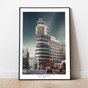 May include: A black and white photograph of a building in Madrid, Spain. The building is called "Edificio Capitol" and has a large sign that says "Cine Capitol" on the side. The building is located on a busy street with cars and people walking by.