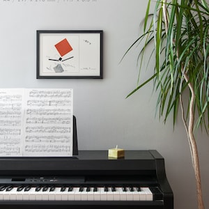 May include: A framed abstract art print with a red square and black lines hangs above a black piano. Sheet music rests on the piano's music stand. A small, square, green candle sits on the piano's surface. A tall green plant is in the background.
