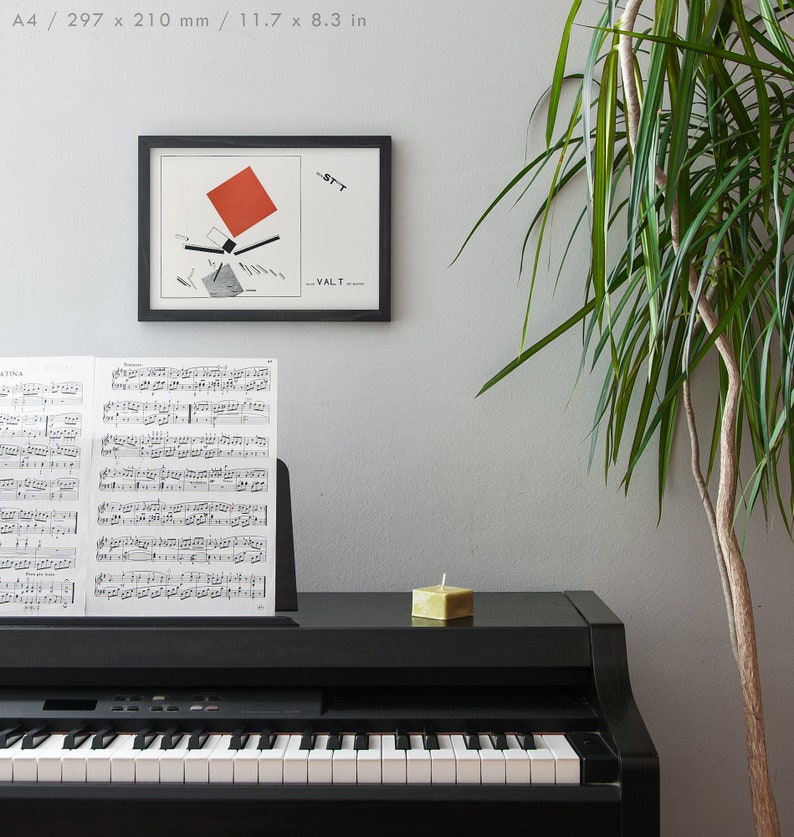 May include: A framed abstract art print with a red square and black lines hangs above a black piano. Sheet music rests on the piano's music stand. A small, square, green candle sits on the piano's surface. A tall green plant is in the background.