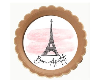 Puede incluir: Un plato de papel marrón con un borde festoneado presenta un círculo blanco con un fondo de acuarela rosa y una ilustración en blanco y negro de la Torre Eiffel. El texto "Bon Appetit!" está escrito en cursiva negra debajo de la torre.