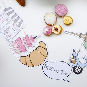 K&ouml;nnte beinhalten: Ein Pariser Partybanner mit Illustrationen einer B&auml;ckerei, eines Kuchens, eines Croissants, eines Rollers, des Eiffelturms und einer Sprechblase mit der Aufschrift "Millie ist Zwei!"