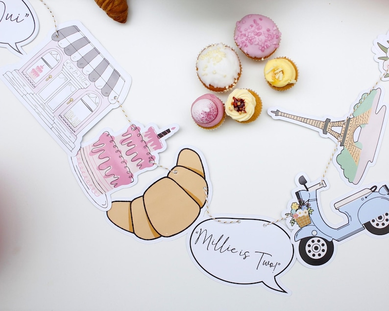 K&ouml;nnte beinhalten: Ein Pariser Partybanner mit Illustrationen einer B&auml;ckerei, eines Kuchens, eines Croissants, eines Rollers, des Eiffelturms und einer Sprechblase mit der Aufschrift "Millie ist Zwei!"