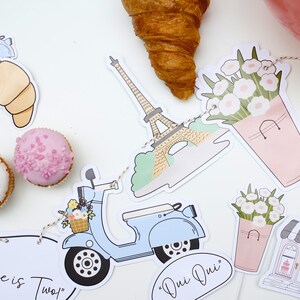 K&ouml;nnte beinhalten: Ein Pariser Partybanner mit Illustrationen eines Rollers, des Eiffelturms, von Croissants, Blumen in Eimern und einer B&auml;ckerei. Das Banner enth&auml;lt den Text "Millie ist Zwei!" und "Oui Oui".