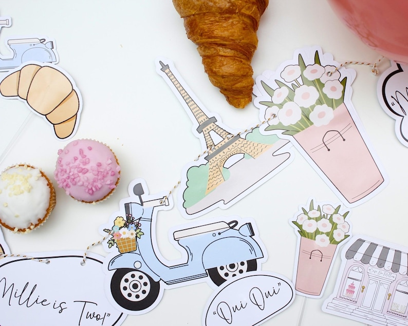 K&ouml;nnte beinhalten: Ein Pariser Partybanner mit Illustrationen eines Rollers, des Eiffelturms, von Croissants, Blumen in Eimern und einer B&auml;ckerei. Das Banner enth&auml;lt den Text "Millie ist Zwei!" und "Oui Oui".