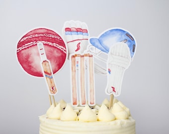 May include: A white frosted cake decorated with cricket-themed cake toppers. The toppers include a red cricket ball, a cricket bat, a blue helmet, a white wicket keeper's pad, and a set of wooden cricket stumps.