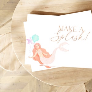 May include: A white card with a pink mermaid holding balloons with a starfish and seashell design. The text "Make a Splash!" is written in cursive on the card.