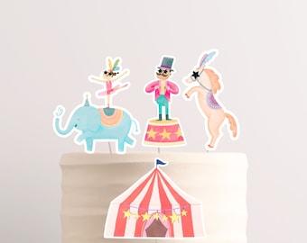 May include: A set of five circus-themed cake toppers. The toppers include a blue elephant with a pink and orange patterned saddle, a girl in a pink tutu balancing on the elephant, a man in a red and green suit standing on a pink and yellow platform, a white horse with a pink and purple patterned saddle, and a red and white striped tent with yellow stars.