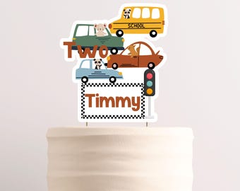 May include: A cake topper featuring a yellow school bus, a blue car, a red car, and a sign that says "Two Timmy". The topper is decorated with animal characters and a traffic light.