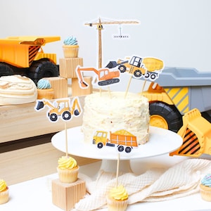 Puede incluir: Un pastel de construcción amarillo y naranja con adornos de grúa, excavadora, camión volquete y bulldozer. El pastel está rodeado de cupcakes y vehículos de construcción de juguete.