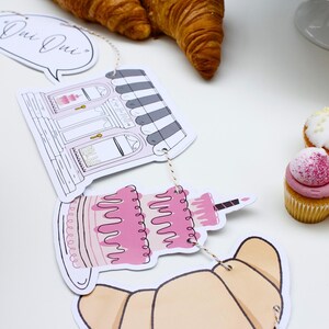 K&ouml;nnte beinhalten: Ein Papierbanner mit Illustrationen einer B&auml;ckerei, eines Kuchens und Croissants. Das Banner sagt "Oui Oui" und ist mit einer Schnur befestigt. Auf einer wei&szlig;en Oberfl&auml;che befinden sich auch Cupcakes mit rosa und wei&szlig;em Frosting.