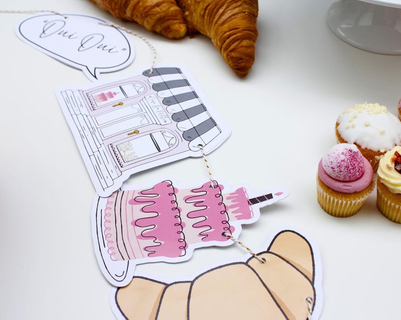 K&ouml;nnte beinhalten: Ein Papierbanner mit Illustrationen einer B&auml;ckerei, eines Kuchens und Croissants. Das Banner sagt "Oui Oui" und ist mit einer Schnur befestigt. Auf einer wei&szlig;en Oberfl&auml;che befinden sich auch Cupcakes mit rosa und wei&szlig;em Frosting.