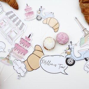 K&ouml;nnte beinhalten: Ein rosa-wei&szlig;es Pariser Partybanner mit dem Text "Millie ist Zwei" und "Oui Oui". Das Banner enth&auml;lt eine B&auml;ckerei, einen Roller, ein Croissant, eine Torte und den Eiffelturm.