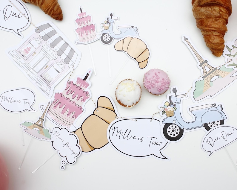 K&ouml;nnte beinhalten: Ein rosa-wei&szlig;es Pariser Partybanner mit dem Text "Millie ist Zwei" und "Oui Oui". Das Banner enth&auml;lt eine B&auml;ckerei, einen Roller, ein Croissant, eine Torte und den Eiffelturm.