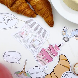 K&ouml;nnte beinhalten: Ein Pariser Partybanner mit Illustrationen einer B&auml;ckerei, einer Torte, eines Rollers, Croissants und des Eiffelturms. Das Banner enth&auml;lt auch Sprechblasen mit dem Text "Oui Oui", "Millie ist Zwei" und "Merci Beaucoup!"