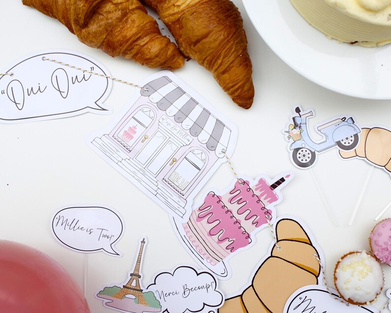 K&ouml;nnte beinhalten: Ein Pariser Partybanner mit Illustrationen einer B&auml;ckerei, einer Torte, eines Rollers, Croissants und des Eiffelturms. Das Banner enth&auml;lt auch Sprechblasen mit dem Text "Oui Oui", "Millie ist Zwei" und "Merci Beaucoup!"