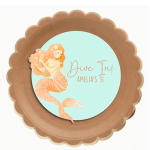 May include: A brown paper party plate with a round, turquoise blue sticker featuring a mermaid with orange hair and tail, holding a seashell. The sticker says "Dive In! Amelia's 31!"