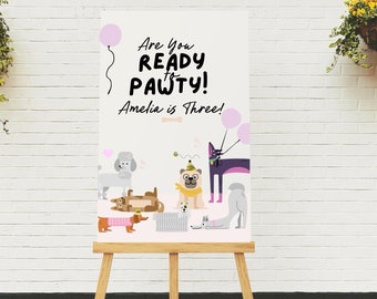 Puede incluir: Un cartel blanco con el texto "Are you ready to pawty! Amelia is Three!" e ilustraciones de perros y un gato. El cartel está en un caballete de madera.