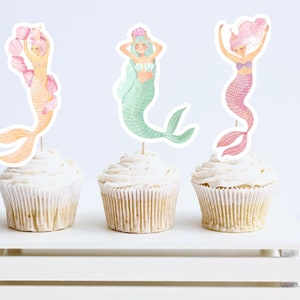 May include: Three cupcakes decorated with mermaid toppers. The toppers are made of paper and feature mermaids with pink, green, and blue tails. The cupcakes are frosted with white frosting.