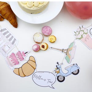 K&ouml;nnte beinhalten: Ein Pariser Partybanner mit Illustrationen einer B&auml;ckerei, eines Kuchens, eines Croissants, eines Rollers, des Eiffelturms und Blumen. Das Banner enth&auml;lt auch den Text "Millie ist Zwei!" und "Merci Beacoup".