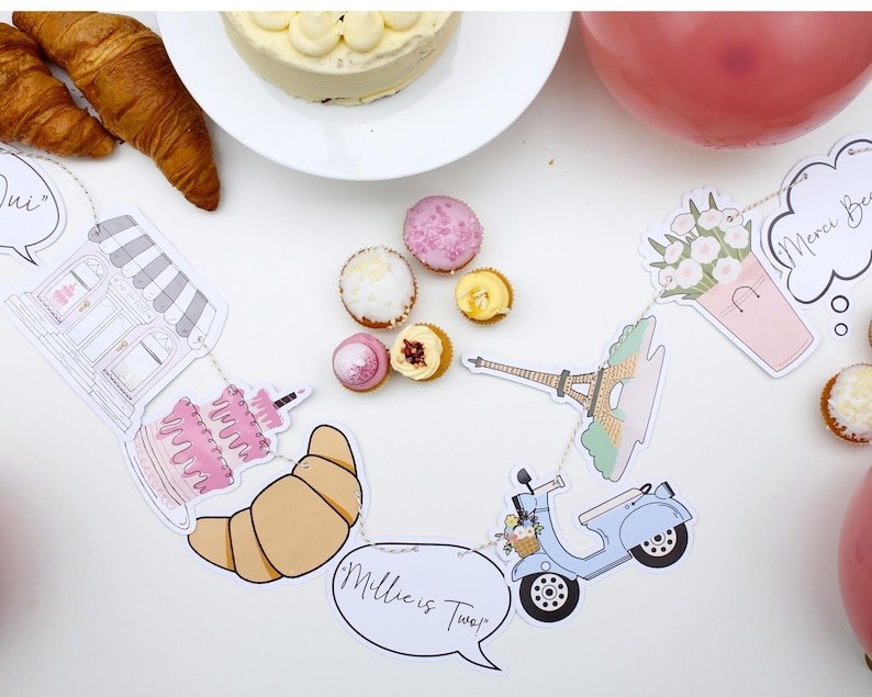 K&ouml;nnte beinhalten: Ein Pariser Partybanner mit Illustrationen einer B&auml;ckerei, eines Kuchens, eines Croissants, eines Rollers, des Eiffelturms und Blumen. Das Banner enth&auml;lt auch den Text "Millie ist Zwei!" und "Merci Beacoup".