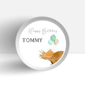 May include: A white round sticker with the text "Happy Birthday TOMMY" and a watercolor illustration of a brown triceratops with green and blue balloons.