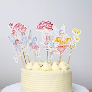 Puede incluir: Un pastel redondo con glaseado amarillo, cubierto con decoraciones de hadas y hongos. Las hadas tienen vestidos coloridos y alas. Los hongos son rojos con puntos blancos. El pastel está sobre un plato blanco.