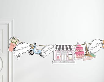 May include: A white string banner with various illustrations and text. The banner includes a pink flower pot, a blue scooter, a speech bubble with the text "Oui Oui", a pink storefront with the text "Cafe en Ville", a pink cake with a tower of the Eiffel Tower, and a speech bubble with the text "Millie is Two".