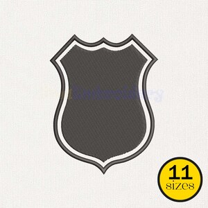 May include: A dark gray shield-shaped embroidery design featuring a thick black border and a thin white inner outline. The emblem has a classic crest silhouette with a pointed bottom and a scalloped top edge, presented on a textured white background.