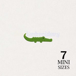 May include: A green embroidered alligator design on a white fabric background. The design features a small, stylized crocodile with a curved tail and a black eye. The text 7 MINI SIZES is printed in black in the lower right corner of the image.