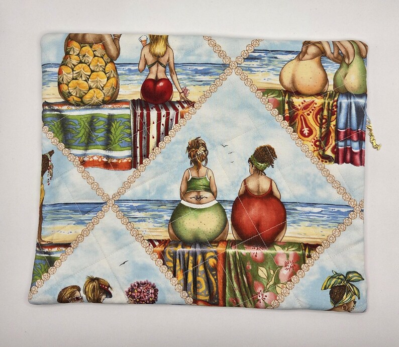 May include: A rectangular quilted fabric pouch featuring a repeating beach scene print. The design depicts multiple figures sitting on towels by the ocean, rendered in vibrant colors including red, green, and yellow. A gold-toned zipper pull is visible.