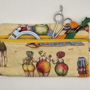 May include: A rectangular quilted fabric pouch with a yellow zipper closure. The pouch features a printed pattern of people in swimsuits on a sandy beach. Inside the pouch are silver metal scissors, a light blue embroidery hoop, and green and orange embroidery floss.