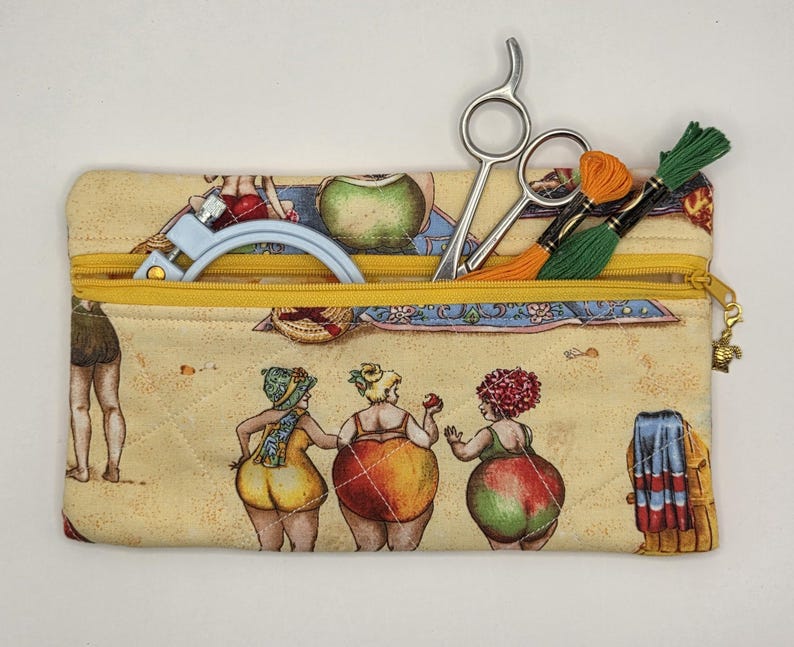May include: A rectangular quilted fabric pouch with a yellow zipper closure. The pouch features a printed pattern of people in swimsuits on a sandy beach. Inside the pouch are silver metal scissors, a light blue embroidery hoop, and green and orange embroidery floss.