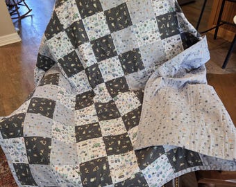 May include: A patchwork quilt featuring a repeating pattern of construction vehicles, including excavators and dump trucks, in shades of gray, light blue, and white. The quilt is constructed from alternating solid-colored and patterned squares.