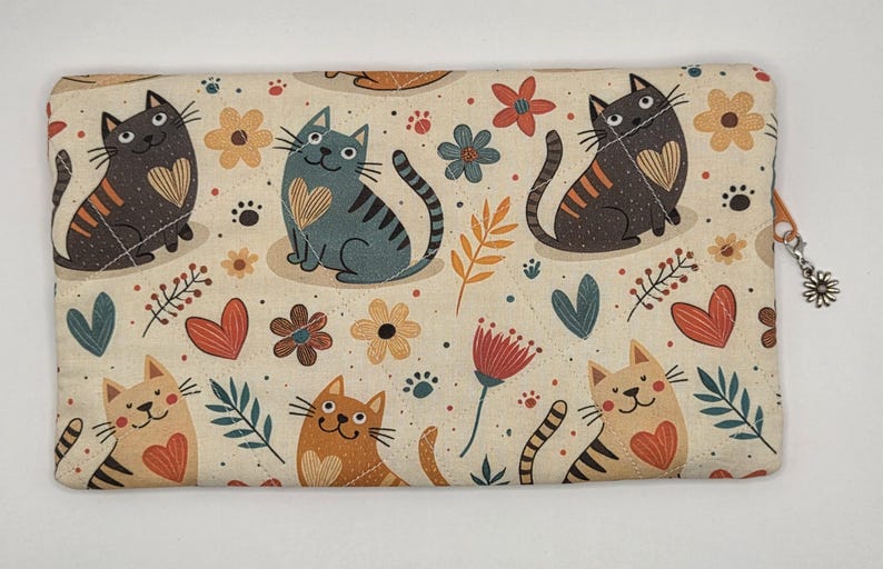 May include: A rectangular fabric pouch featuring a cream background with a repeating pattern of cats in various colors, including black, gray, orange, and tan. Each cat has a heart on its chest. The design includes small flowers, leaves, and hearts.