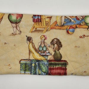 May include: A rectangular quilted zipper pouch featuring a tan background with a beach scene print. The design includes illustrations of people relaxing on the sand, beach chairs, and a ball. A small gold-toned turtle charm is attached to the zipper.