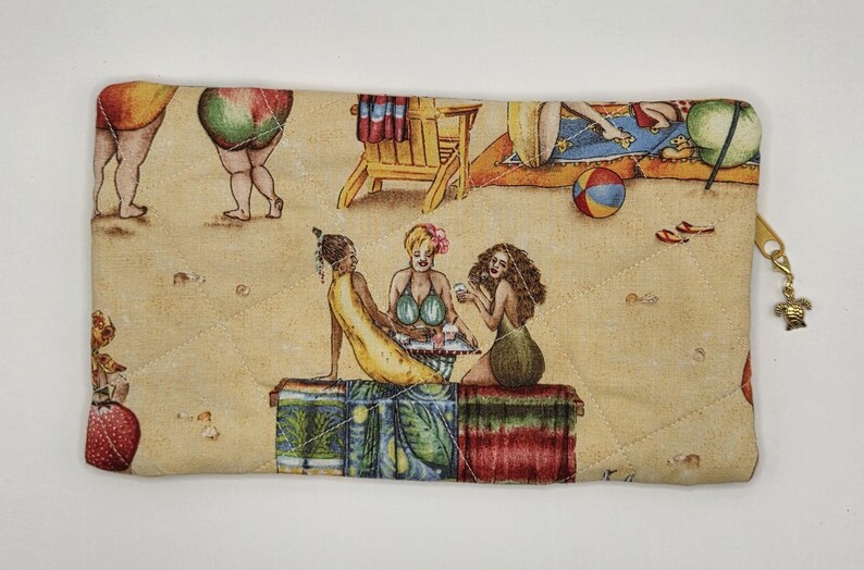 May include: A rectangular quilted zipper pouch featuring a tan background with a beach scene print. The design includes illustrations of people relaxing on the sand, beach chairs, and a ball. A small gold-toned turtle charm is attached to the zipper.