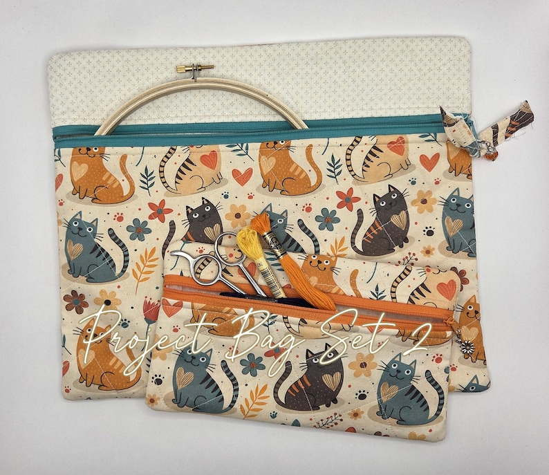 May include: A set of two fabric project bags featuring a repeating pattern of orange and gray cats with hearts and flowers. The larger bag has a teal zipper and contains a wooden embroidery hoop. The smaller bag has an orange zipper and holds scissors and embroidery floss.