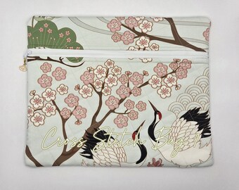 May include: A rectangular quilted zipper pouch featuring a light blue background with a pattern of pink cherry blossoms, green pine branches, and two white cranes with black wing tips. The text Cross Stitch Bag 1 is printed in gold script.